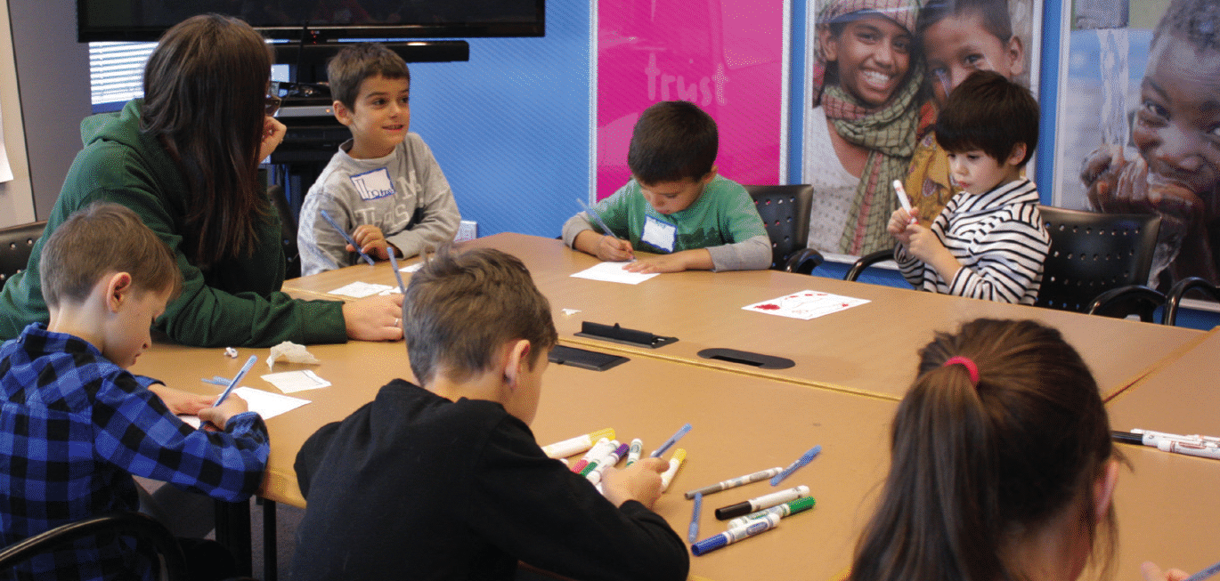 A group of young children sits around a table, engaged in drawing and coloring activities. An adult sits with them, guiding their work. The room has colorful posters in the background, with images of smiling children and words like 'Trust.' The setting appears to be a welcoming, educational space designed for youth engagement and development.